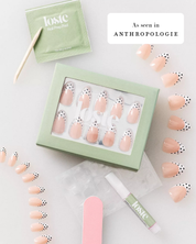 A flat lay of a Loste press-on nail kit on a white background, featuring a mint green box of peach-colored nails with black polka dots, a packet of nail prep, a wooden tool, a pink nail file, and a lip treatment tube. Text overlays read 'As seen in Anthropologie'.