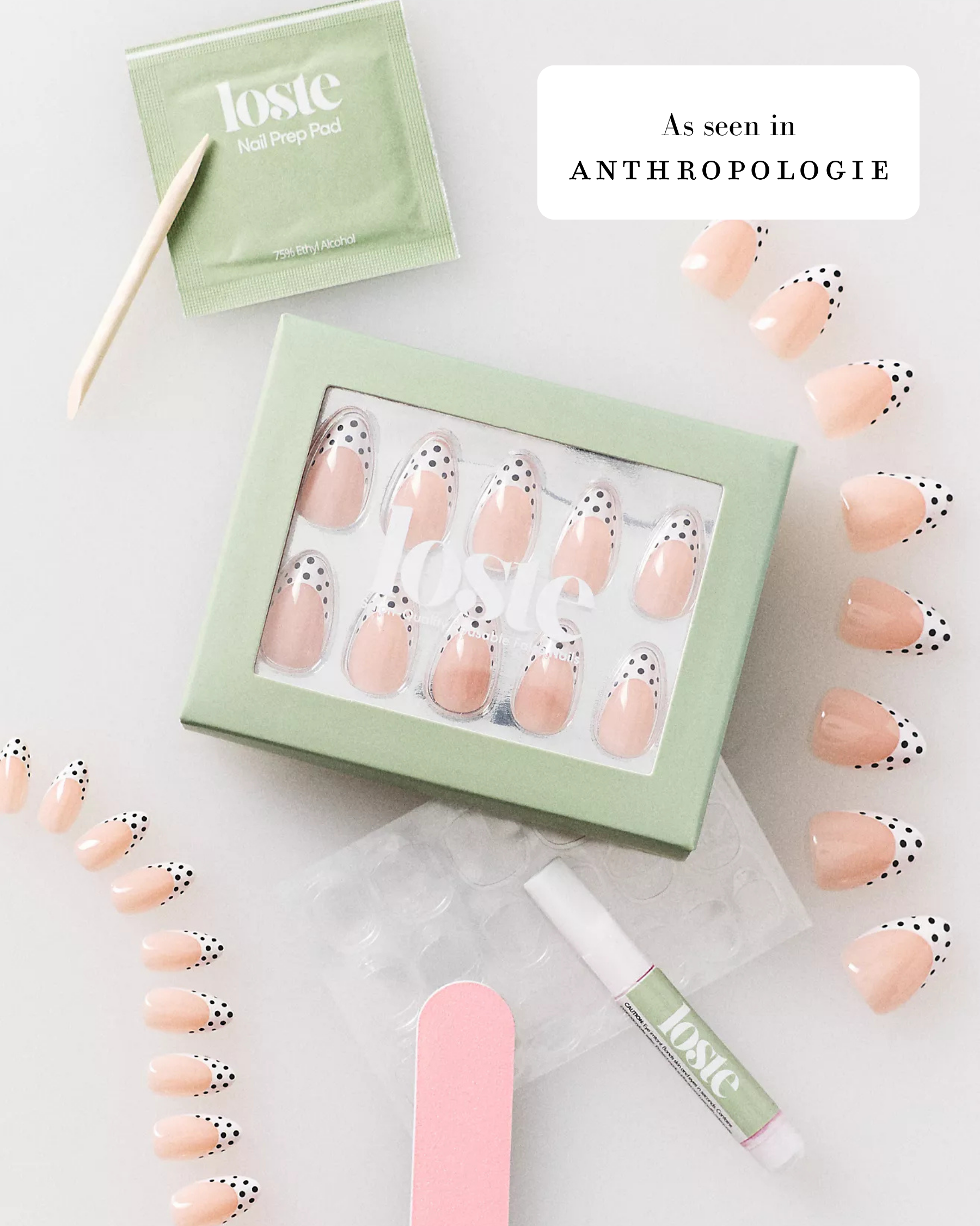 A flat lay of a Loste press-on nail kit on a white background, featuring a mint green box of peach-colored nails with black polka dots, a packet of nail prep, a wooden tool, a pink nail file, and a lip treatment tube. Text overlays read 'As seen in Anthropologie'.