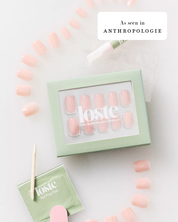 A styled flat-lay of a Loste false nail set in a pale mint green box, with several nude pink false nails arranged around it on a white background. A small green packet, wooden stick, and nail file are also visible, along with an 'As seen in ANTHROPOLOGIE' badge.