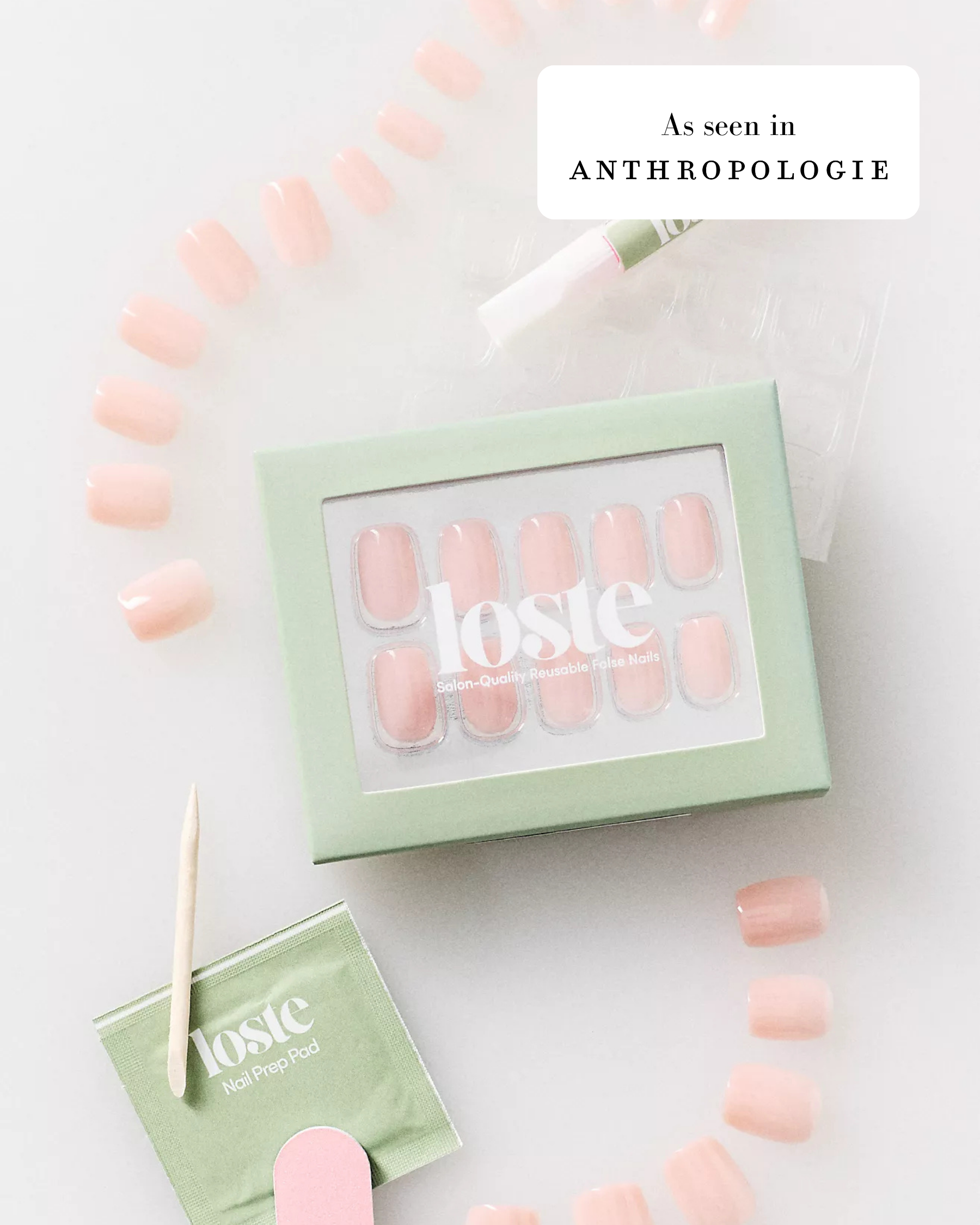 A styled flat-lay of a Loste false nail set in a pale mint green box, with several nude pink false nails arranged around it on a white background. A small green packet, wooden stick, and nail file are also visible, along with an 'As seen in ANTHROPOLOGIE' badge.