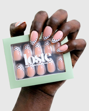 A hand holding a mint green box of Loste press-on nails. The nails have a glossy pale pink base with white French tips and black polka dots.