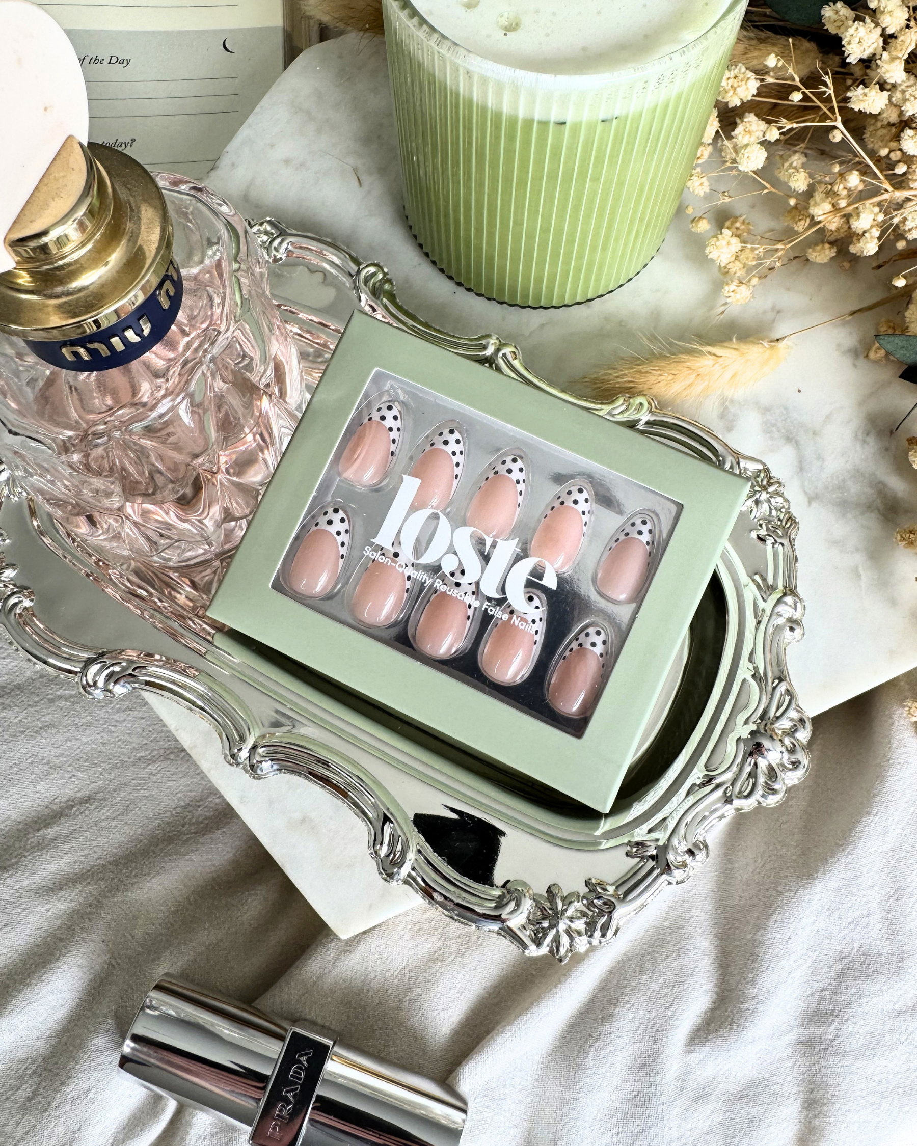 A flat lay with matcha, perfume and flowers and a box of Loste press-on nails in a black and white polka dot design in the middle of the image