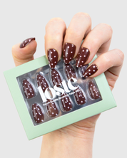 A hand holding a mint green box of loste press-on nails. The nails are glossy dark brown with pink polka dots.