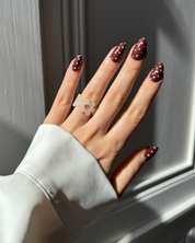 A close-up of a hand with dark brown Loste press-on nails decorated with pink polka dots, wearing a translucent white flower-shaped ring, set against a grey panelled background.
