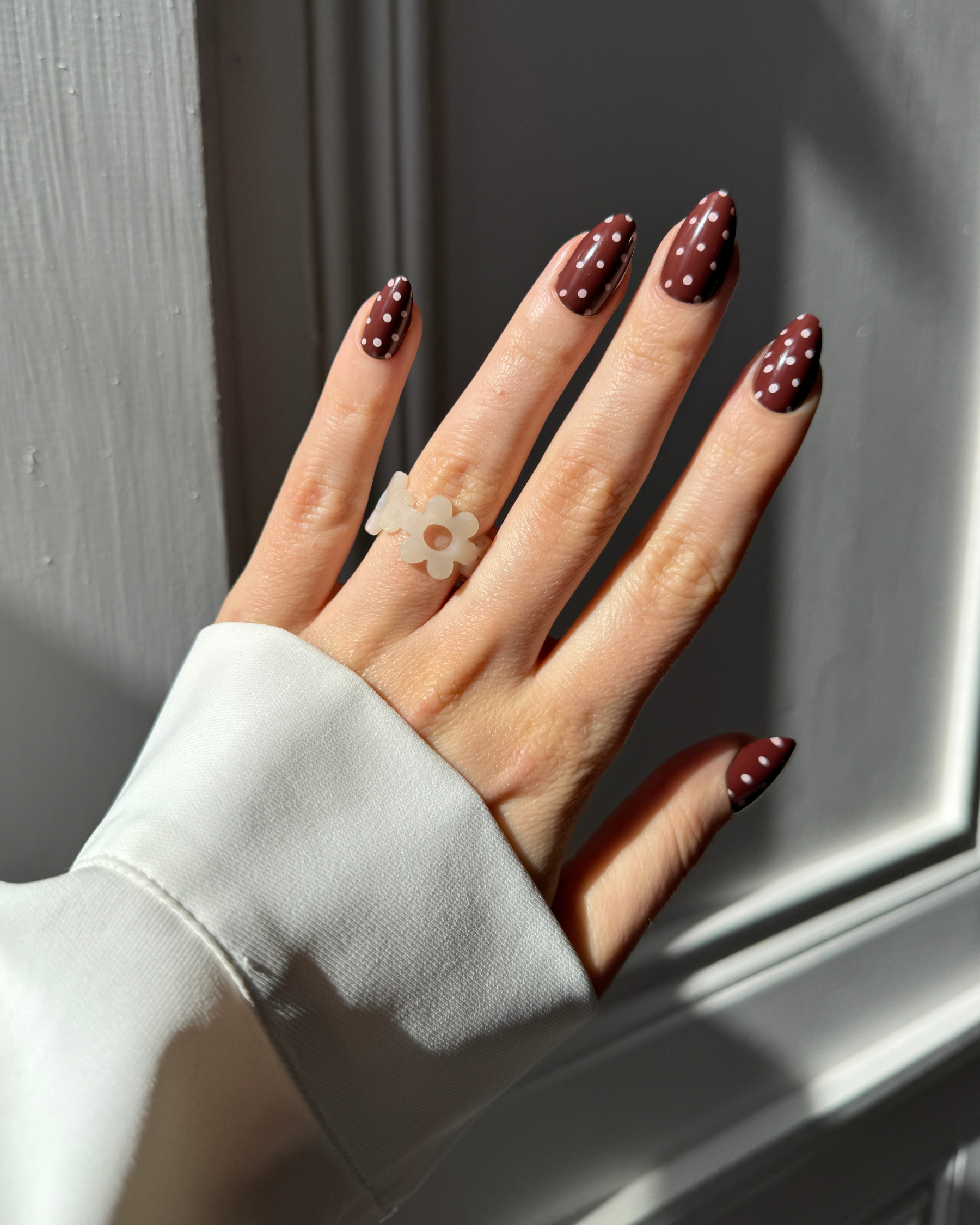 A close-up of a hand with dark brown Loste press-on nails decorated with pink polka dots, wearing a translucent white flower-shaped ring, set against a grey panelled background.
