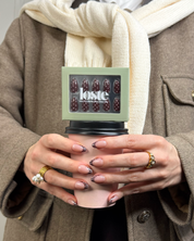 A person wearing a beige coat and a cream knit scarf holds a pink takeaway coffee cup with a black lid. The cup has a small square package on top featuring dark brown polka-dot press-on nails by Loste.