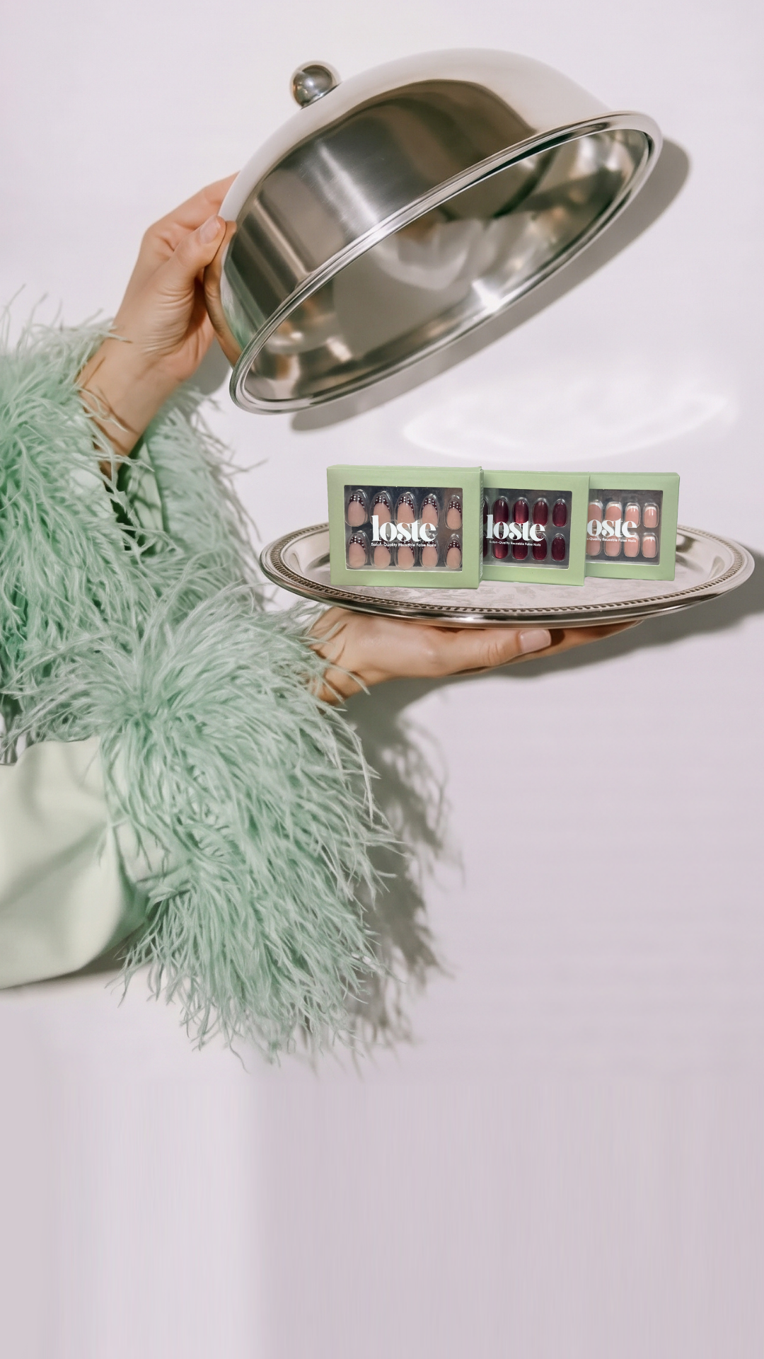 A person in a light green feathered jacket holds a silver cloche dome over a silver tray, revealing three boxes of Loste press-on nails.