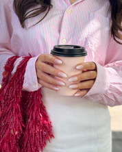 A close-up of a person holding a beige paper coffee cup with a black lid, wearing a pink and white striped button-up shirt and a white skirt. A shiny red tassel bag is draped over one shoulder. Both hands are holding the coffee cup, and she is wearing a set of Loste reusable press-on nails in a long French melt design.