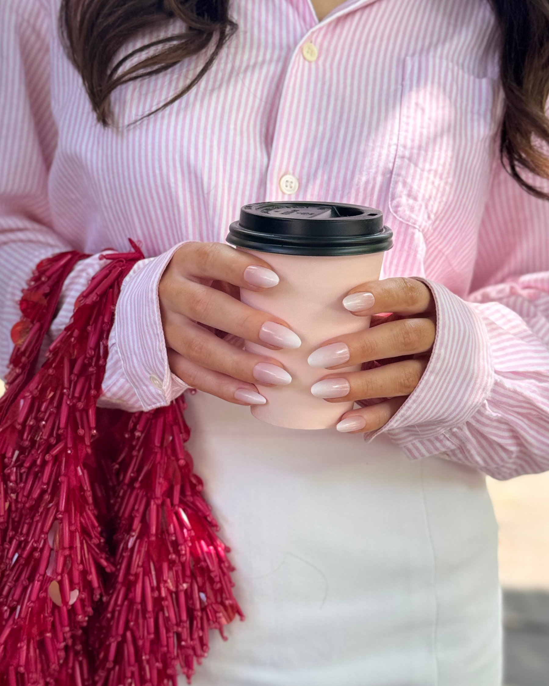A close-up of a person holding a beige paper coffee cup with a black lid, wearing a pink and white striped button-up shirt and a white skirt. A shiny red tassel bag is draped over one shoulder. Both hands are holding the coffee cup, and she is wearing a set of Loste reusable press-on nails in a long French melt design.