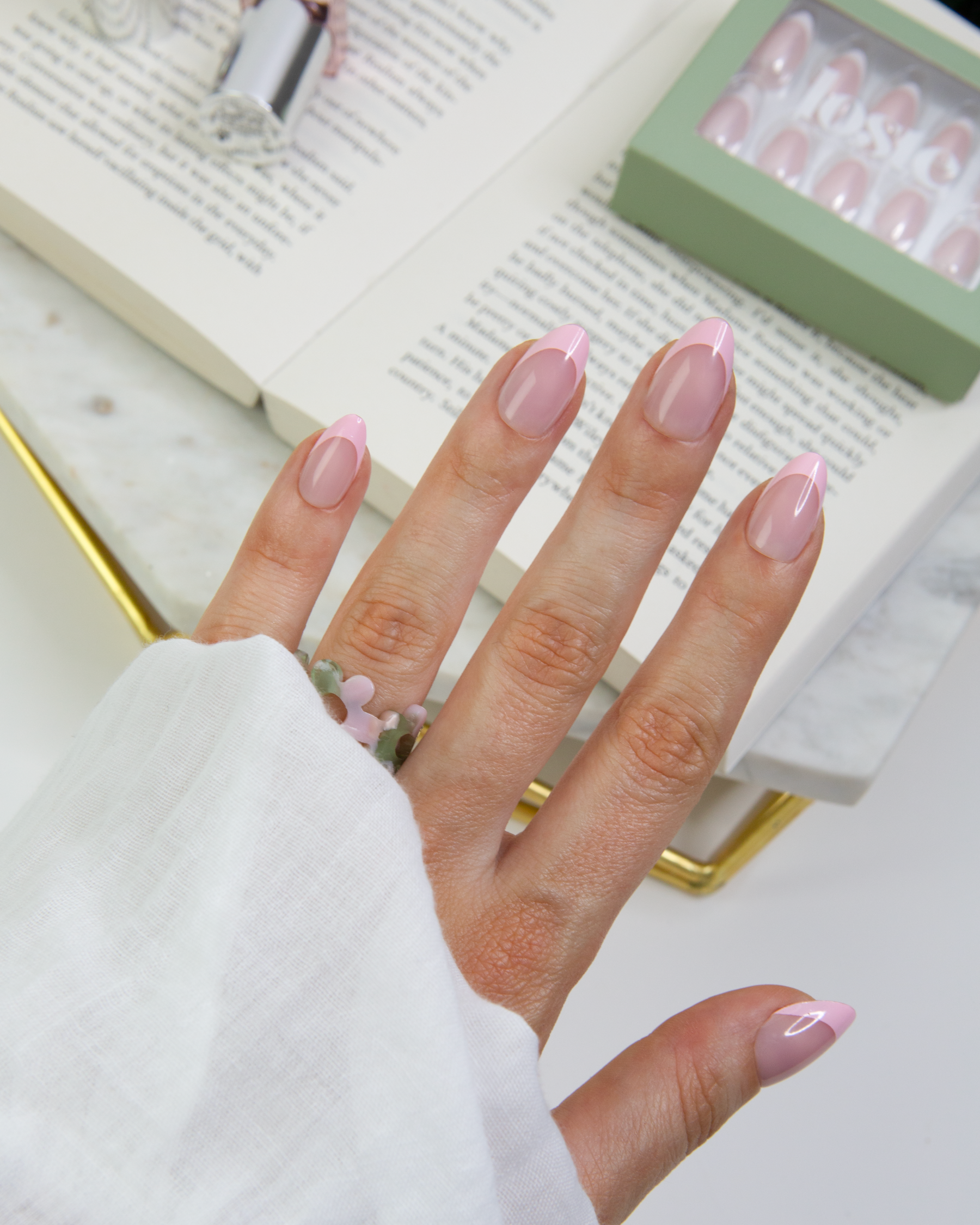 Hand wearing a set of pink French Loste press-on nails over a flatlay include a book and box of Loste nails 