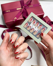 A hand holding a box of Loste false nails and wearing Loste false nails featuring red French nails with a gold and glitter design 