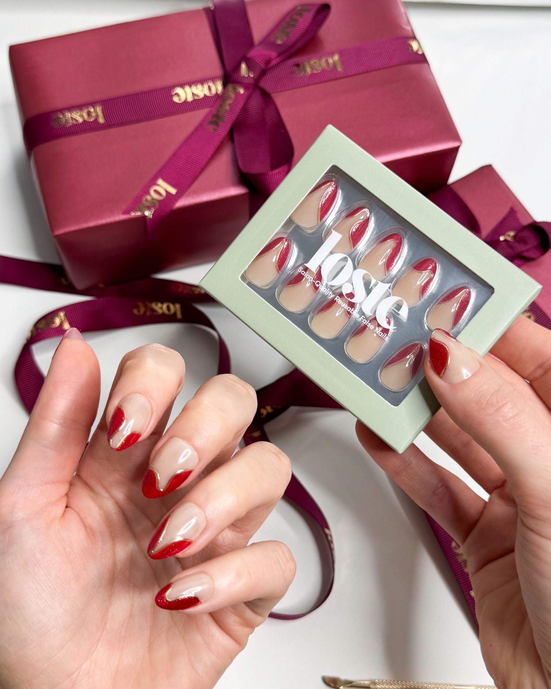 A hand holding a box of Loste false nails and wearing Loste false nails featuring red French nails with a gold and glitter design 
