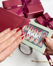 A hand holding a box of Loste false nails and wearing Loste false nails featuring red French nails with a gold and glitter design 