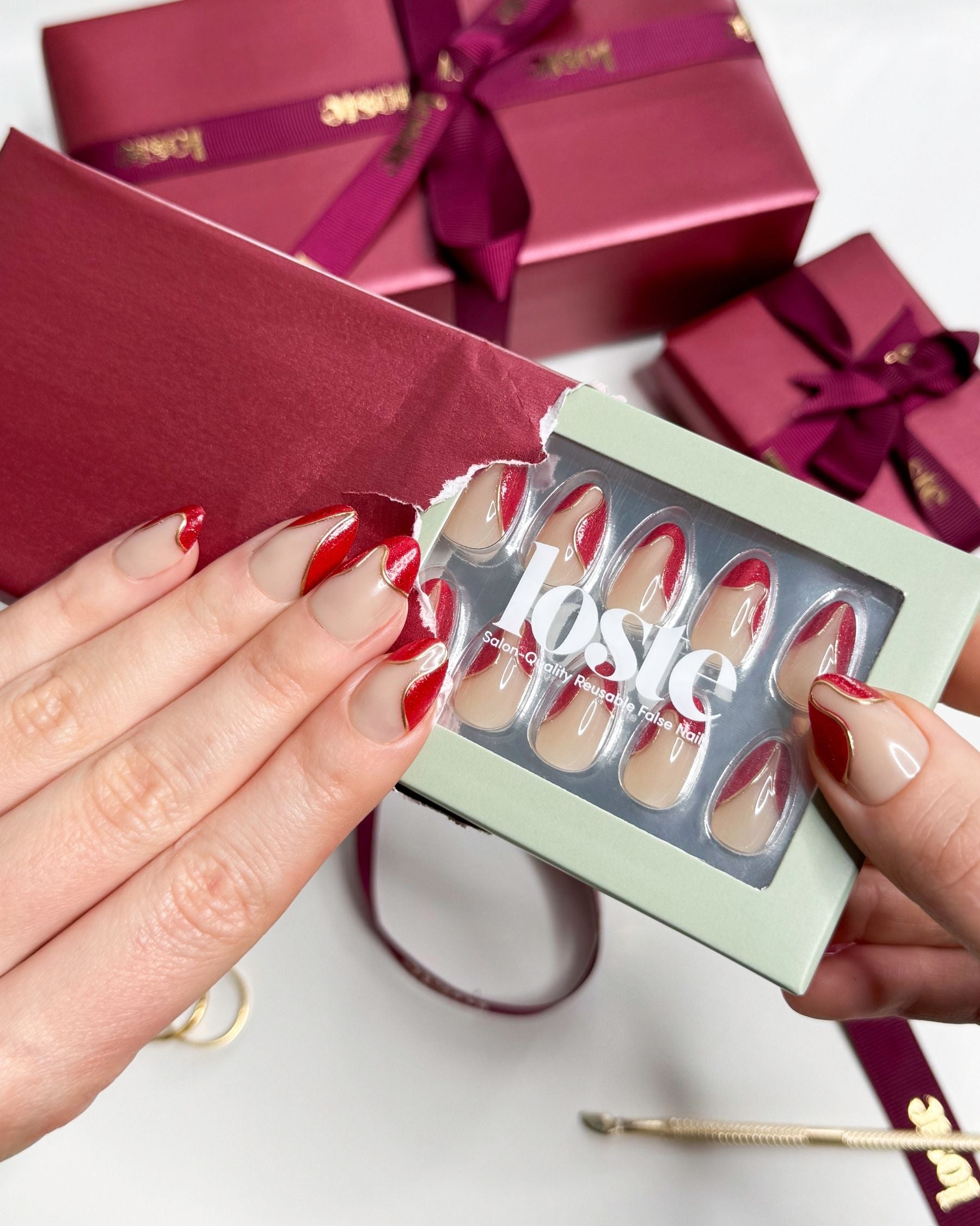 A hand holding a box of Loste false nails and wearing Loste false nails featuring red French nails with a gold and glitter design 