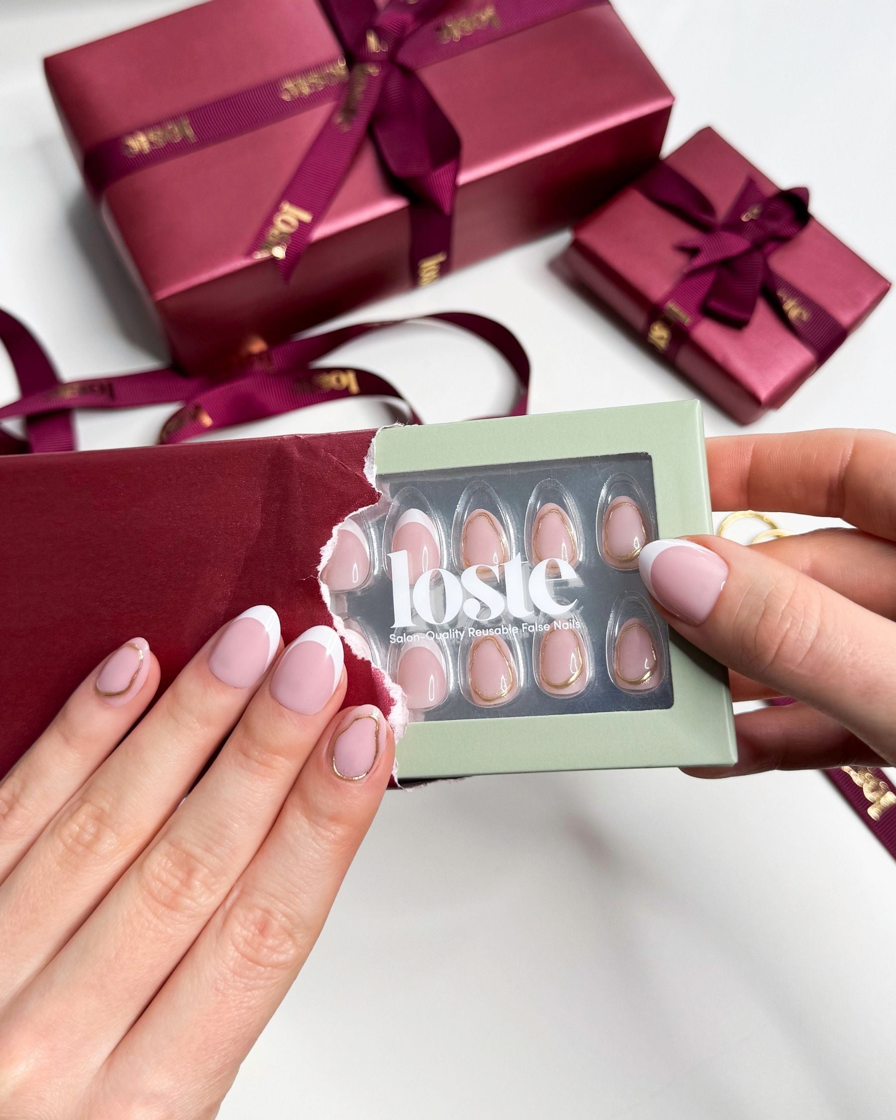 A close-up of hands holding a Loste press-on nail set in a light sage green box, with a torn red paper sleeve revealing the short almond shaped nails. The nails have a pale pink base with a metallic gold accent.