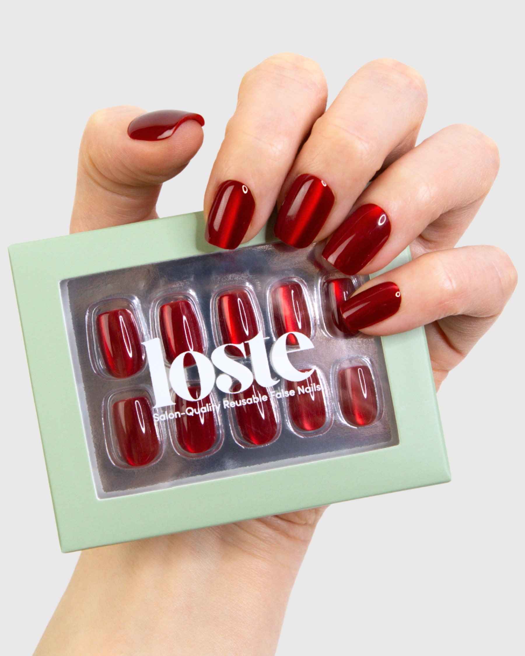 A hand holding a package of Loste red cat-eye press-on nails. The nails have a glossy, deep red colour with a subtle cat-eye reflective sheen, and the hand is shown against a plain light gray background.