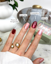 A close-up of a hand displaying five different Loste press-on nails with  name labels: 'Financially Irresponsible' (deep burgundy), 'Sugar Baby' (pale pink), 'Cosmopolitan' (shimmery metallic rose gold), 'Ballerina' (shimmery pale pink), and a box of press-on nails in the background. The hand is also wearing two gold rings with pearl details.