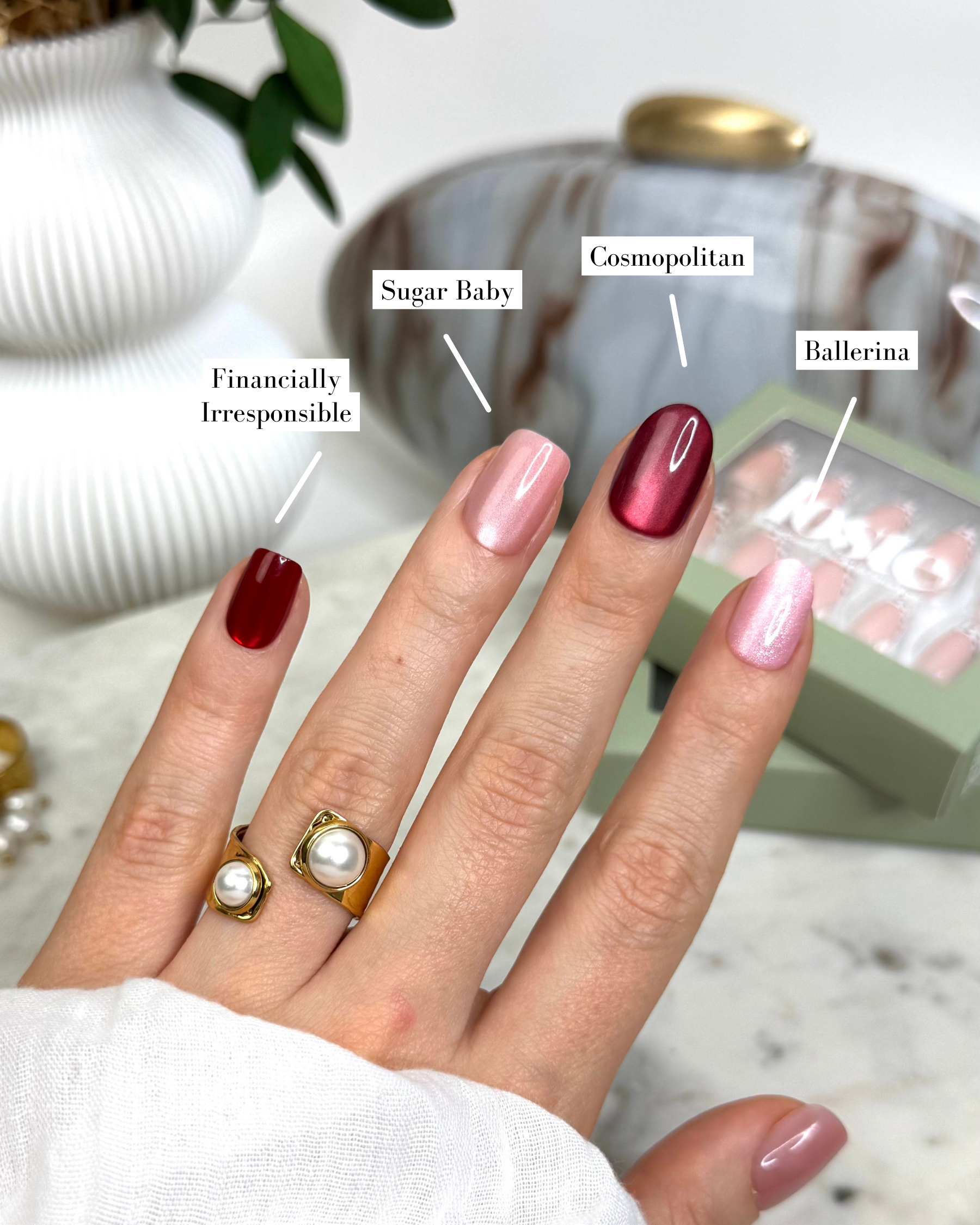 A close-up of a hand displaying five different Loste press-on nails with  name labels: 'Financially Irresponsible' (deep burgundy), 'Sugar Baby' (pale pink), 'Cosmopolitan' (shimmery metallic rose gold), 'Ballerina' (shimmery pale pink), and a box of press-on nails in the background. The hand is also wearing two gold rings with pearl details.