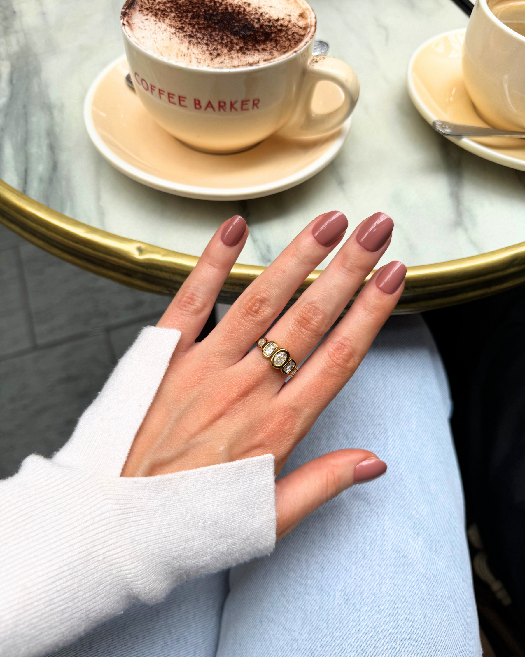  a flatlay showing a coffee table with a coffee cup and a hand wearing a set of Loste press-on nails in a super short brown design